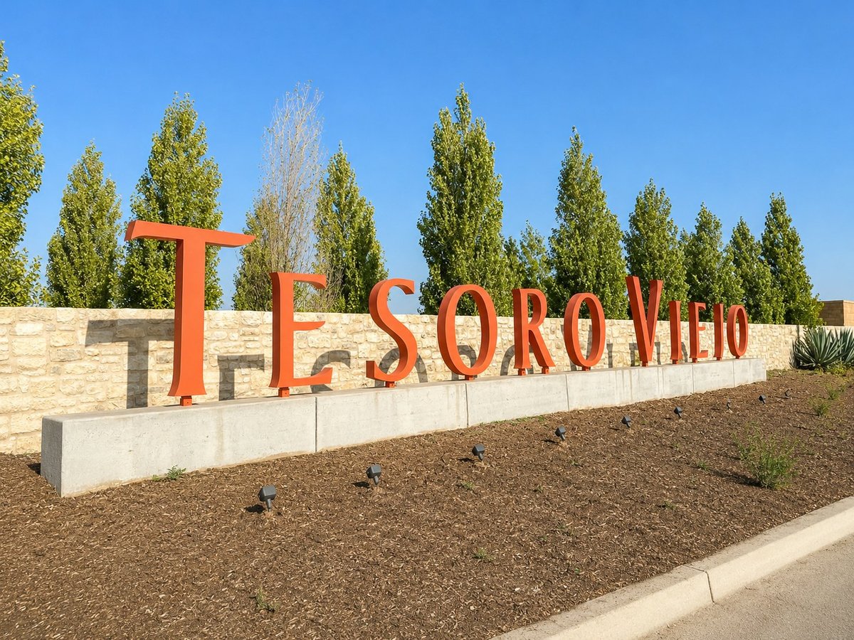 Large orange metal letters spelling TESORO VIEJO sign in a landscaped area with tall green trees and clear blue sky